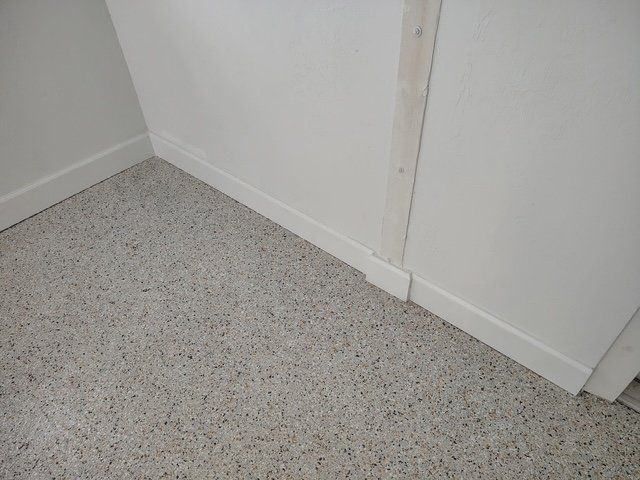 Corner of a white-walled room showing a speckled beige terrazzo-like floor, white baseboards, and a vertical wooden strip anchored to the wall.