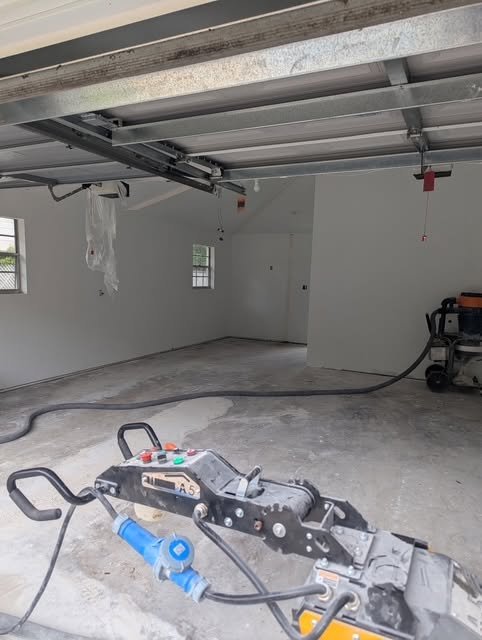 Partially finished garage with exposed garage door tracks, white walls, and a dusty concrete floor; a vacuum hose and power tool are in the foreground.